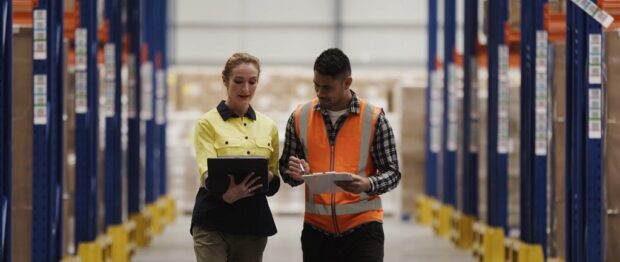 Warehouse workers discussing logistics with clipboards between tall blue storage racks