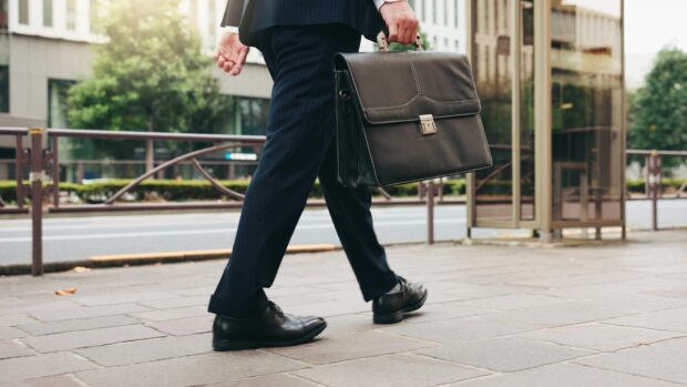 A person in a business suit walks with a black leather briefcase on a city street