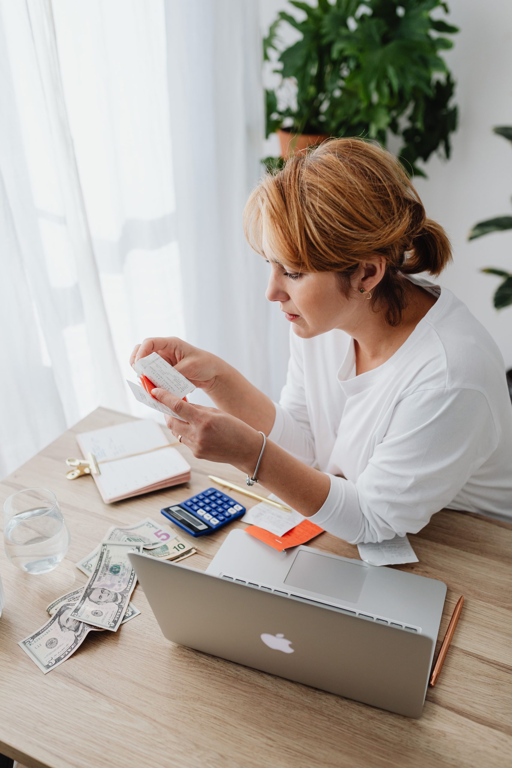 Woman calculating a monthly entertainment budget with laptop, cash, and receipts at desk